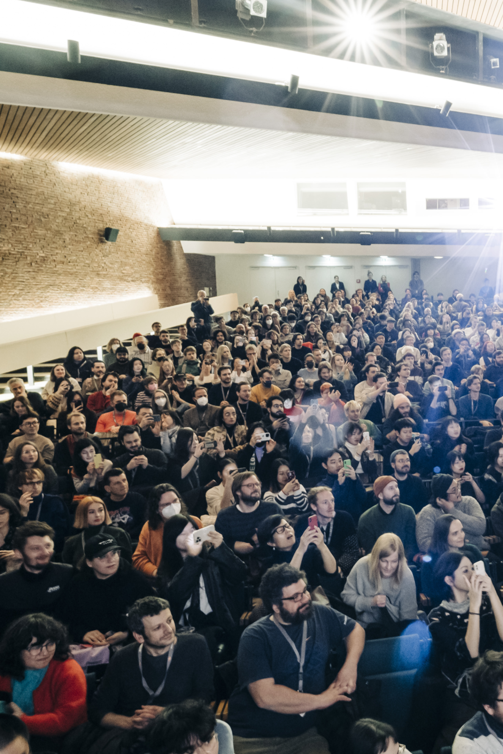 Frontalansicht von Menschen, die in einem Kinosaal sitzen. Das Saallicht ist an, die Reihen sind gefüllt.