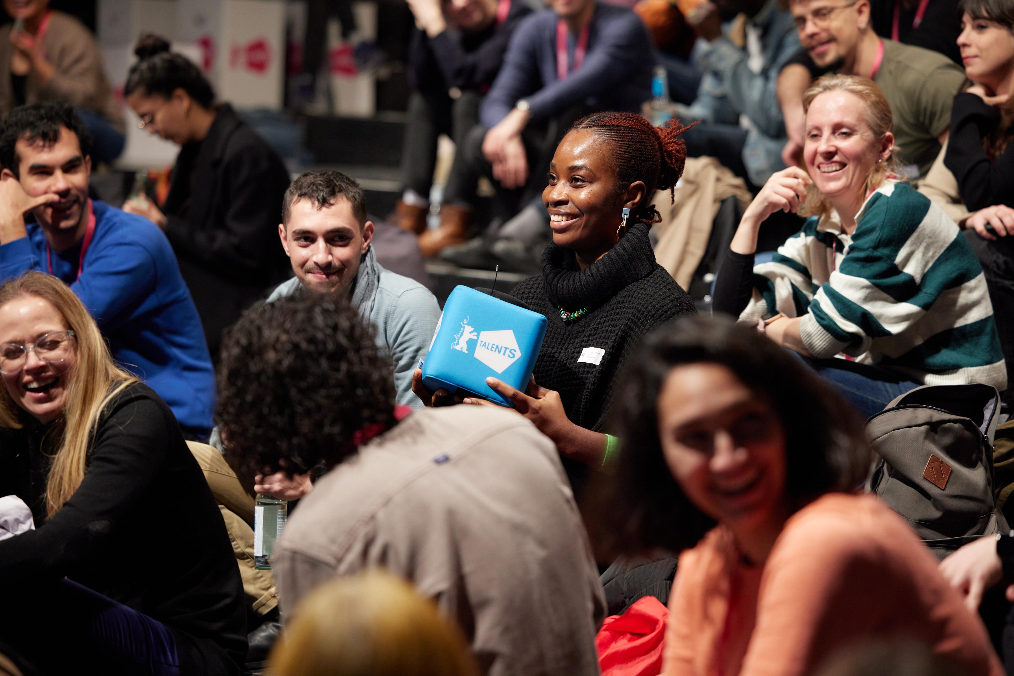 Several young people are sitting on chairs. One person in the middle is holding a blue, cube-shaped loudspeaker with "Berlinale Talents" printed on it.