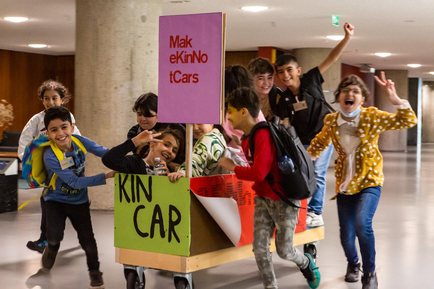 Laughing children push a wooden cart with the lettering "Kin Car" through the HKW building. Children are sitting in the car.
